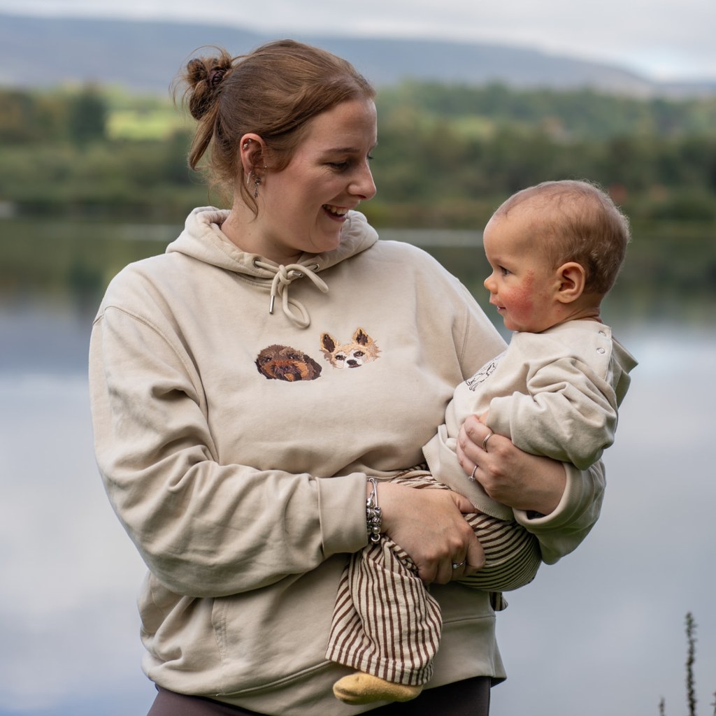 Customer in a cream hoodie embroidered with her two dogs, holding her baby